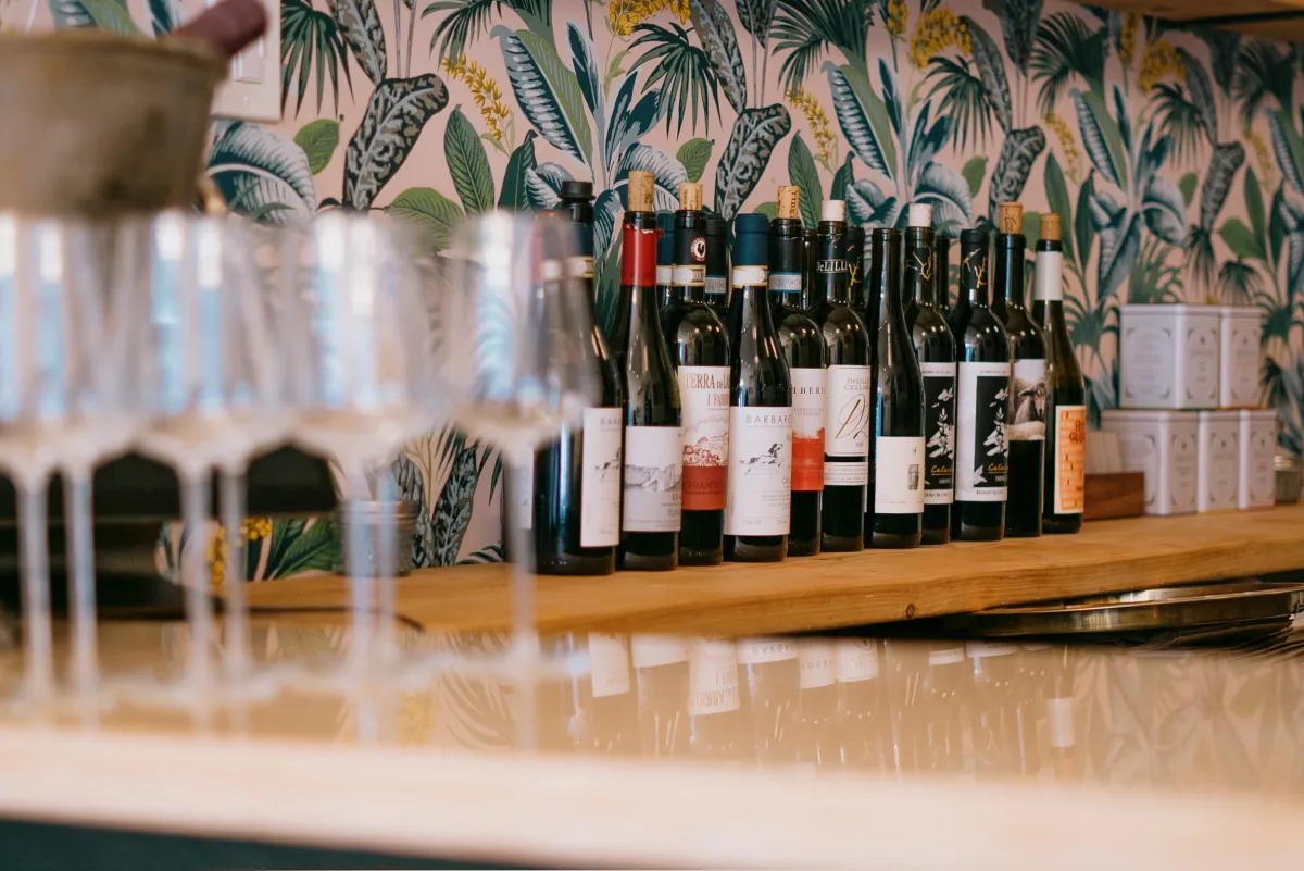 A row of wine bottles lined up on a wooden shelf against a vibrant tropical-leaf wallpaper, with out-of-focus wine glasses in the foreground.