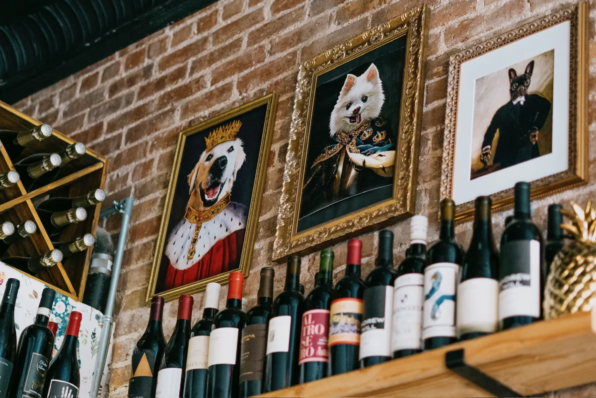 Framed oil paintings of dogs dressed in royal and formal attire hanging on an exposed brick wall above a wooden shelf lined with wine bottles.