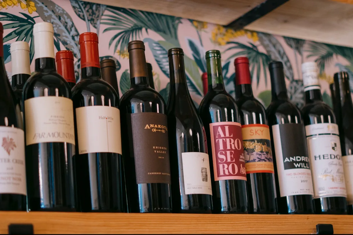 A close-up shot of multiple wine bottles lined up on a wooden shelf against a colorful botanical wallpaper background with pink and green tropical leaves.