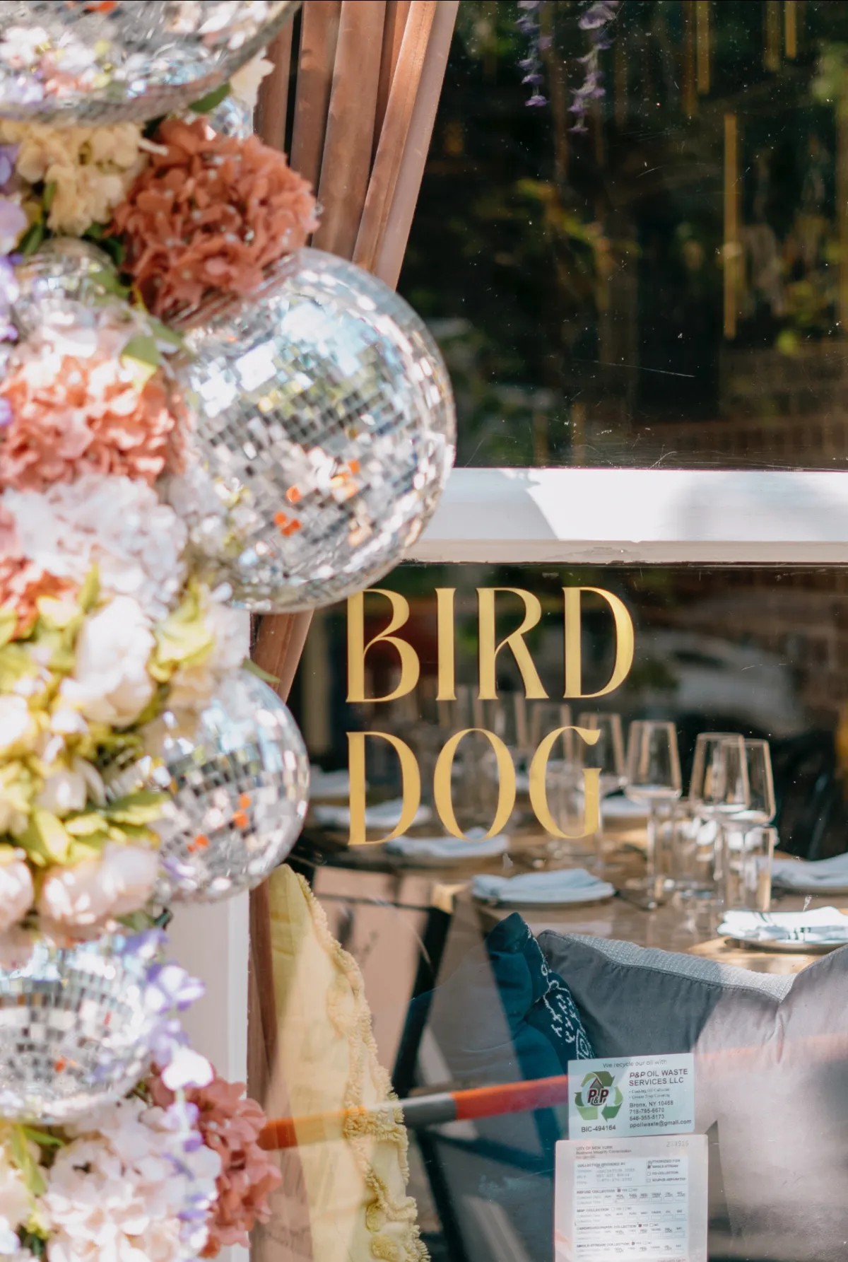 A close-up, angled view of a restaurant window displaying the gold lettering "BIRD DOG," with a decorative floral arrangement and sparkling disco balls hanging along the side frame, and a set dining table visible through the glass.