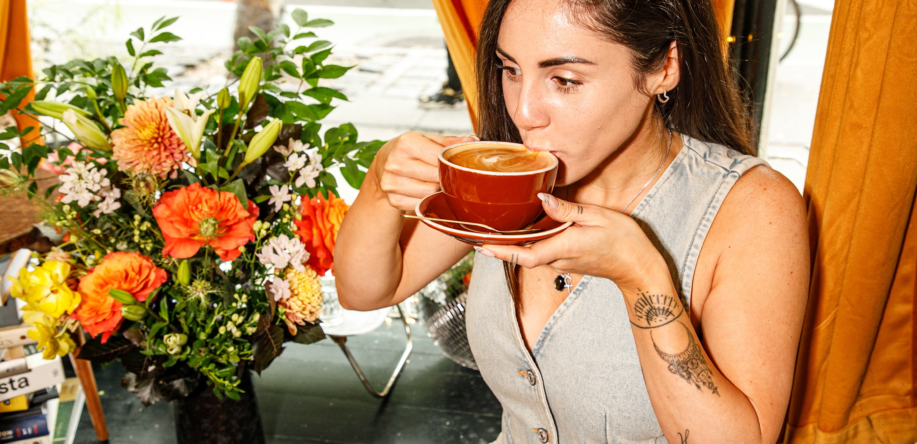 A woman with tattoos on her arm and hand takes a sip of coffee from a brown cup and saucer, sitting next to a large, vibrant floral arrangement with bright orange and yellow flowers.