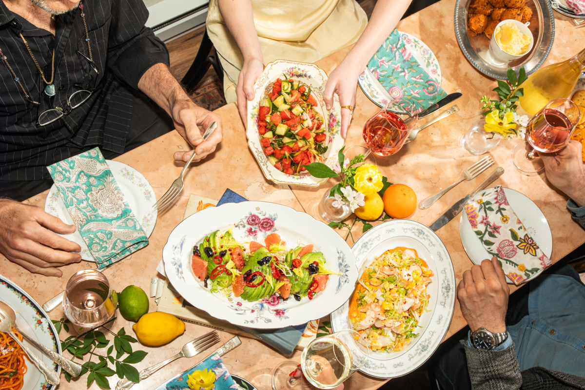 Top-down view of a dinner table filled with colorful salads, pasta dishes, and drinks, with multiple people reaching in to share food.