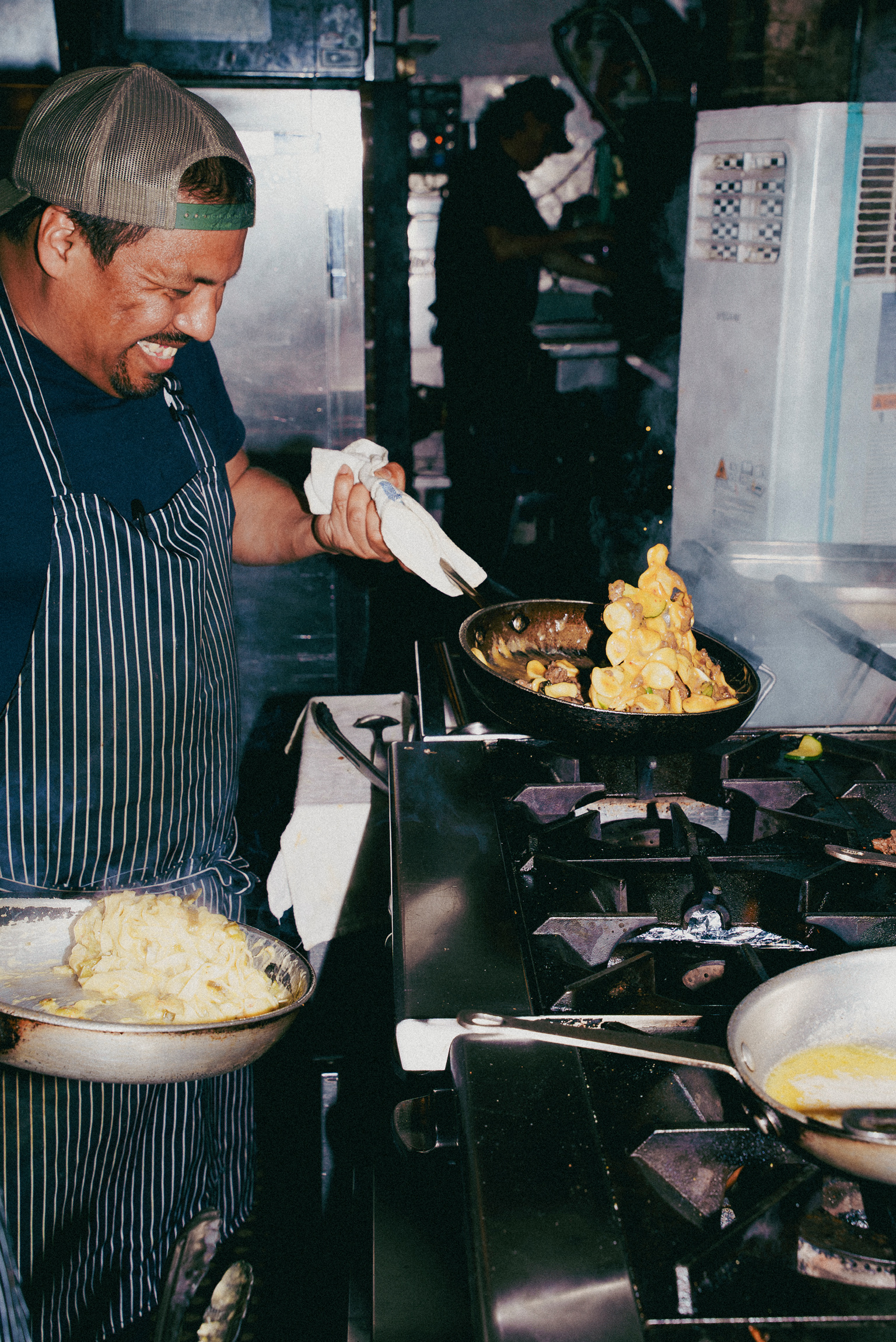 A smiling chef wearing a backward cap and a blue-and-white striped apron stands in a busy commercial kitchen, holding a pan of food while looking down at a skillet on the stove where pasta and vegetables are being tossed.