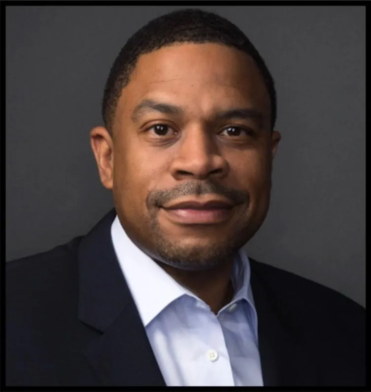 Professional headshot of Reginald Bean. A man with a light beard, wearing a dark suit, light blue shirt, and no tie, against a grey backdrop.