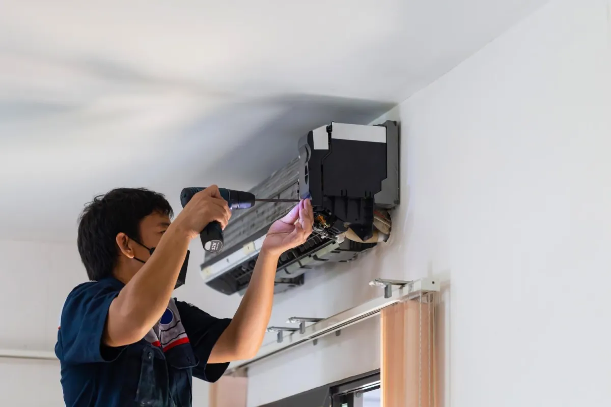 A man works on fixing an air conditioner inside a room, surrounded by tools and equipment necessary for the task.