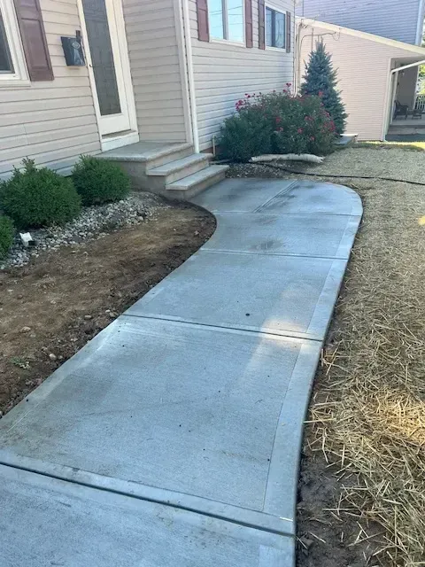 Newly constructed curved concrete walkway leading to the front steps of a house with landscaping in progress, featuring bushes and freshly planted flowers alongside the path.