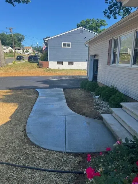 Curved concrete walkway leading from the front steps of a house to the driveway, with surrounding landscaping under development, featuring bushes and freshly planted flowers. A blue house and neighborhood street are visible in the background.