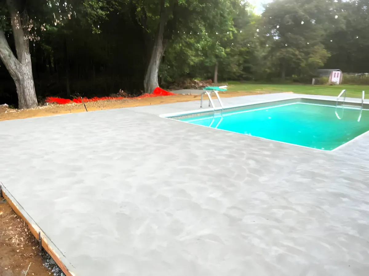 Freshly poured concrete patio surrounding a backyard swimming pool. The area is bordered by trees and greenery, with a small storage shed visible in the background. The pool ladder and diving board are positioned on the right side of the pool.