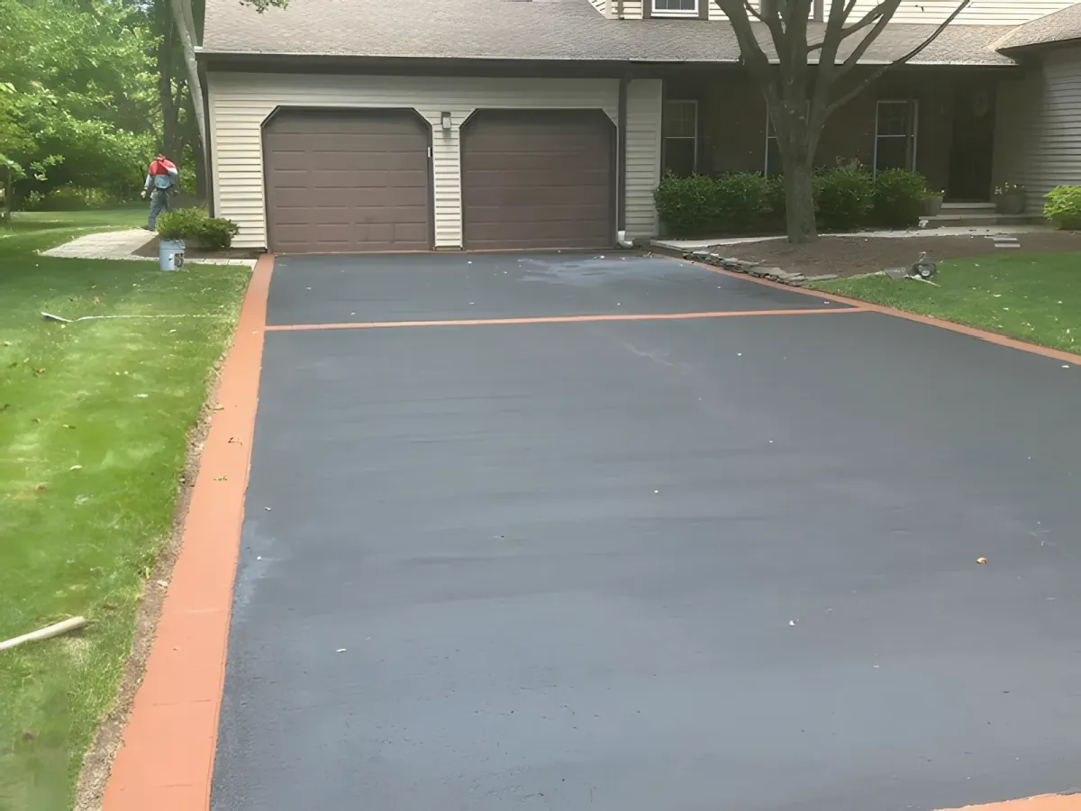 Freshly paved asphalt driveway in front of a two-car garage, bordered by a red edge. A worker is seen in the background near the side of the house with trees and greenery surrounding the area.