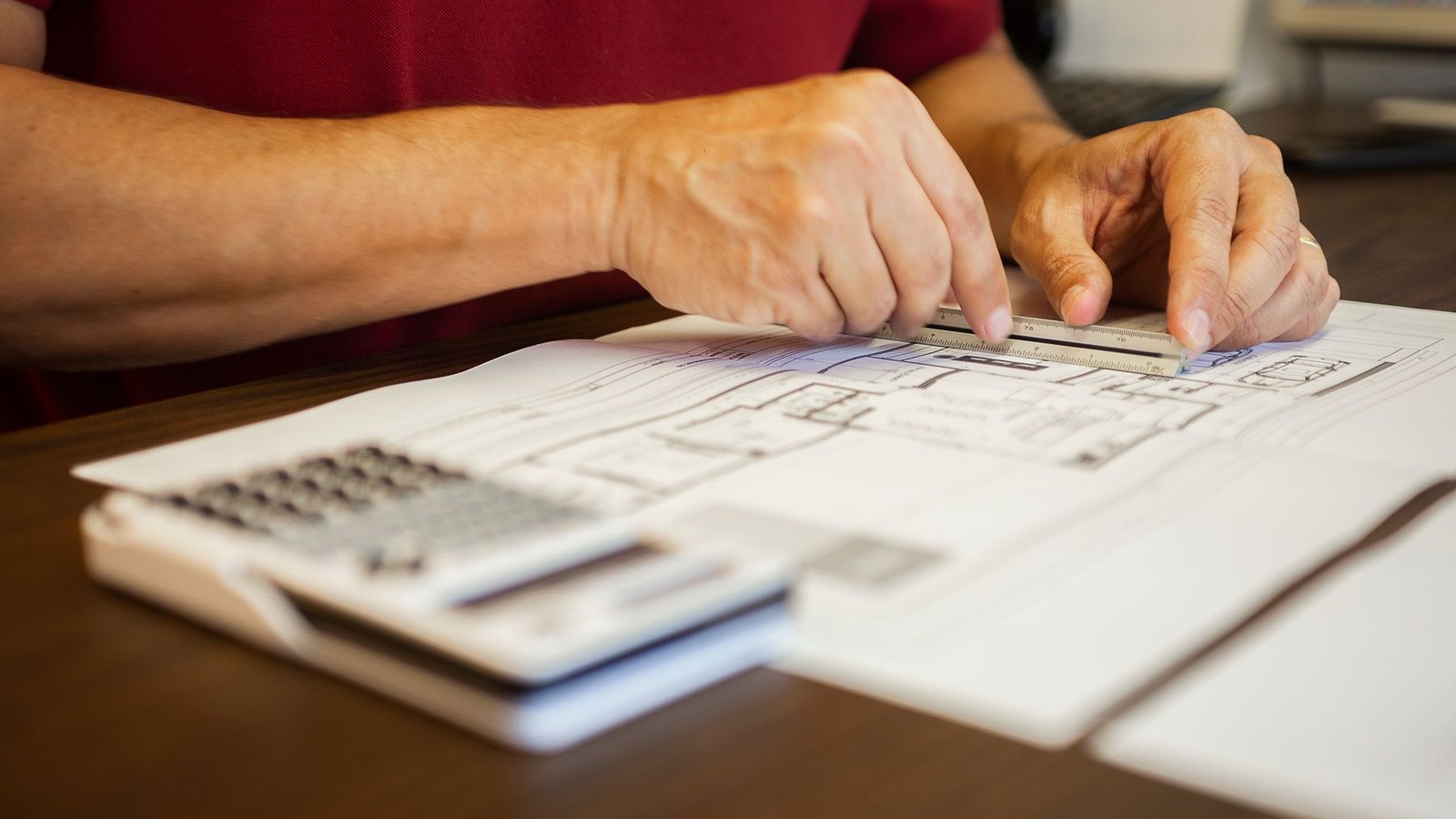 Hands using a scale ruler over a residential floor plan drawing next to a calculator on a desk