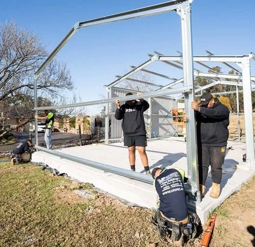 Construction team assembling a custom steel shed frame on a concrete foundation, showcasing licensed builders at work in a residential setting, emphasizing quality and expertise in steel building construction.