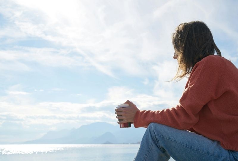 A woman sitting alone by the beach looking out at the water.