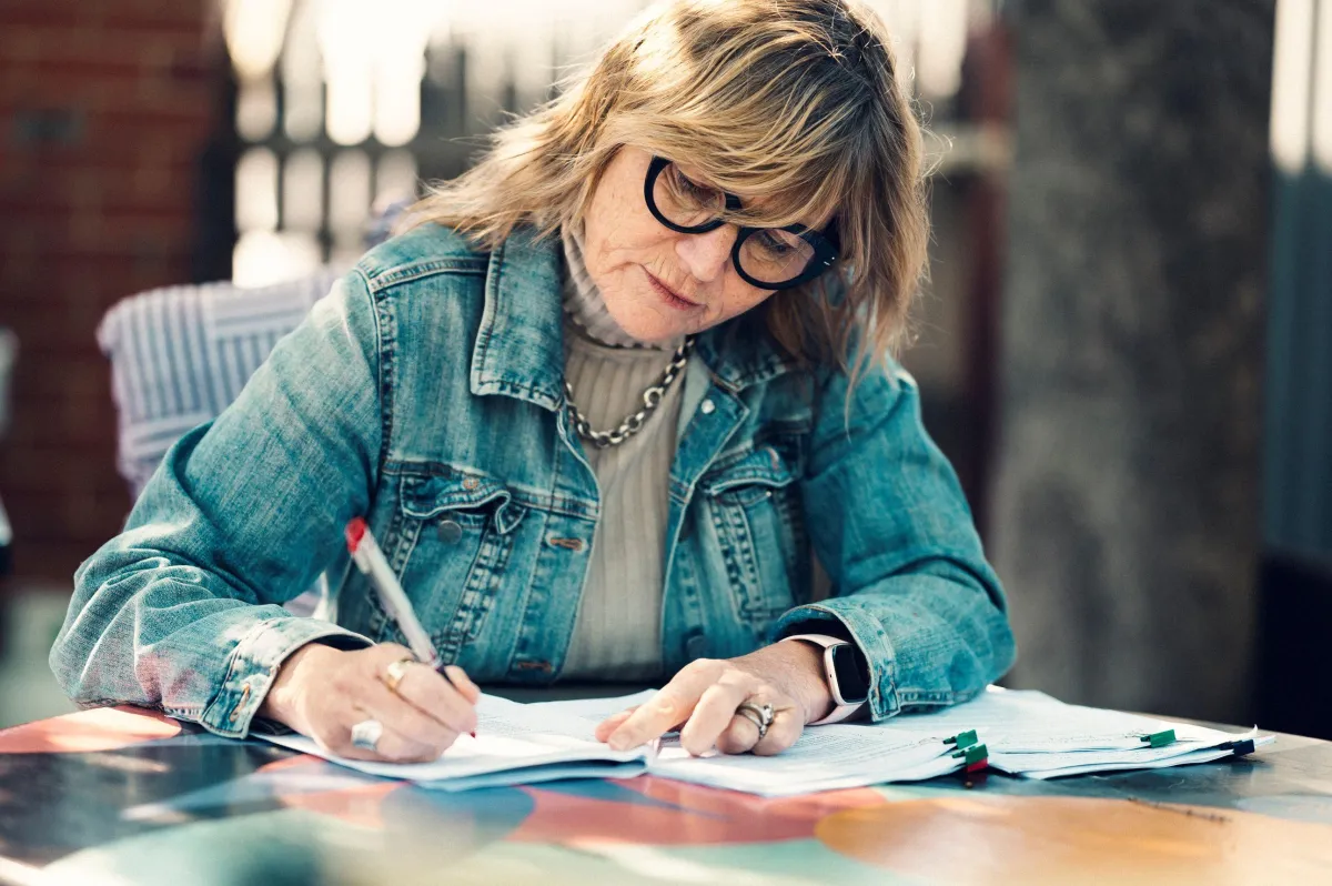 Gry looking thoughtful while sitting at a table outside writing,