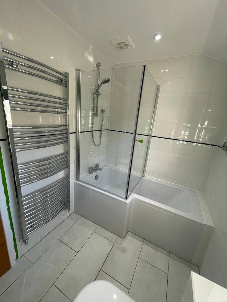 White-tiled bathroom with glass screen shower-over-bath and chrome heated towel rail