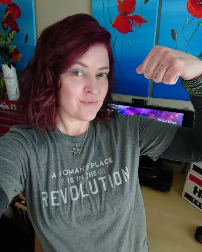 photo of a woman with her fist raised, wearing a t-shirt that says, "A woman's place is in the revolution."