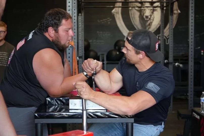 Two competitors engaged in an arm wrestling match at a fitness event, showcasing strength and determination, with gym equipment and spectators in the background.