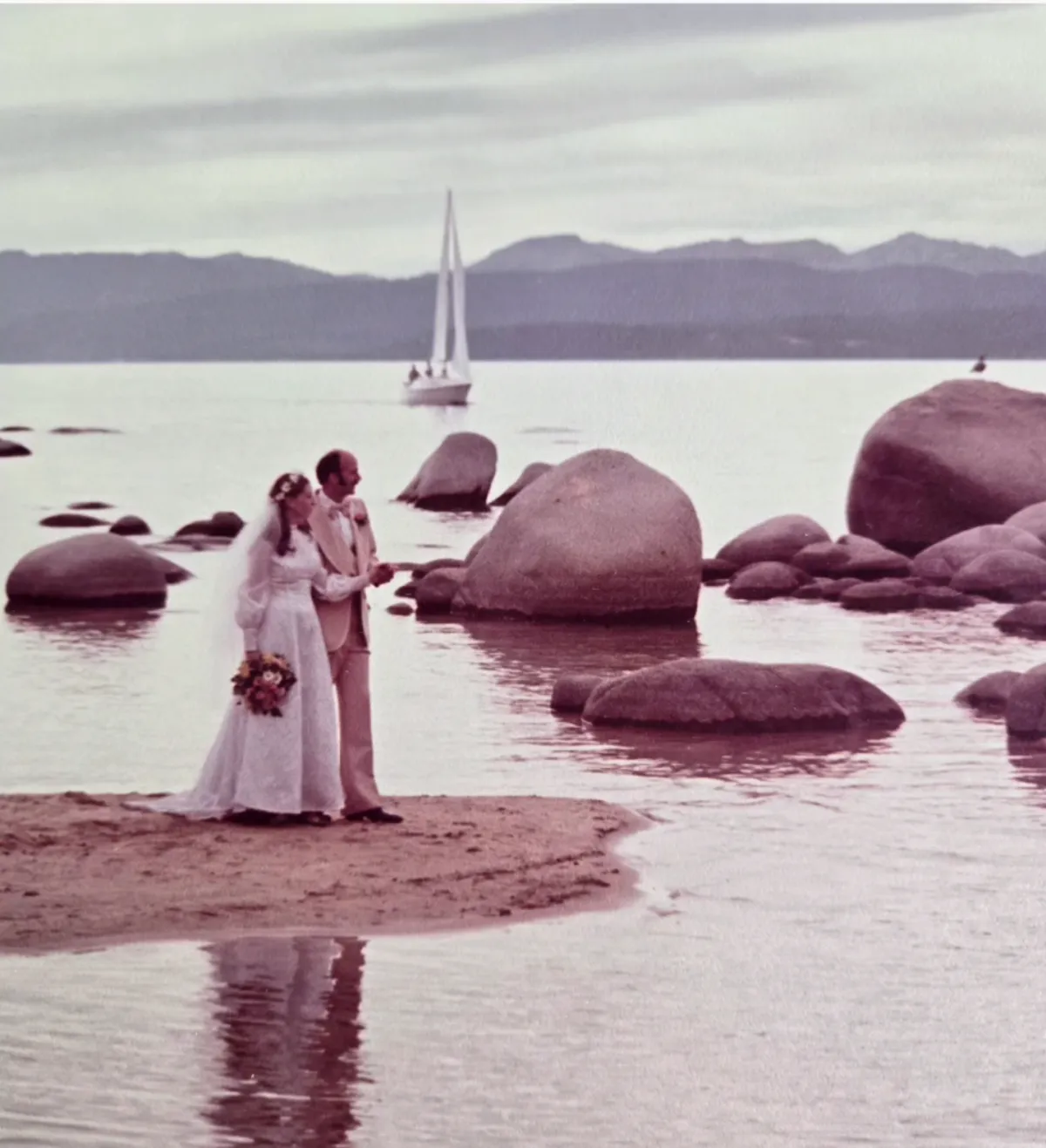 C.S. Fitzpatrick and husband standing on a sandy edge of Sand Harbor State Park, Lake Tahoe with various-sized boulders and a sailboat as the backdrop.