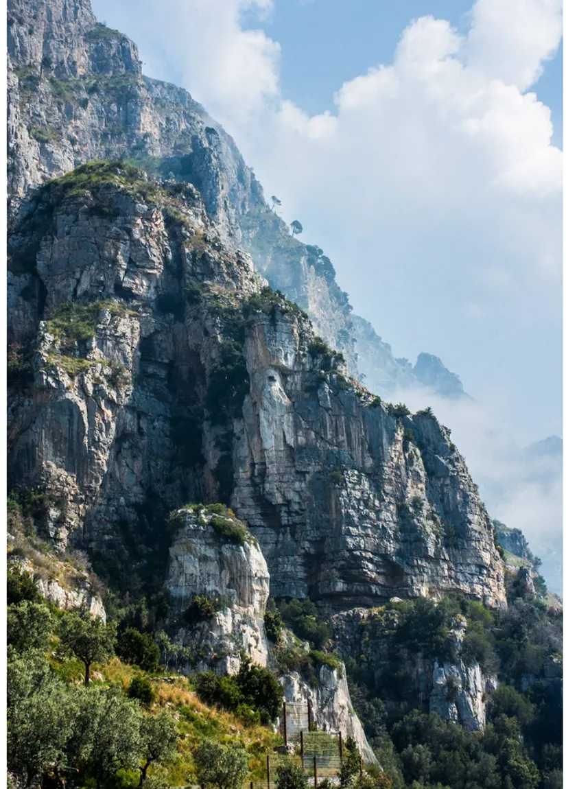 Tall, sheer, and multi-tiered cliff face comprised of seemingly crumbly stone which would be difficult to climb, but greenery still that appears clings to the tops of the cliffs and ominous clouds tuck in between the level of cliff faces.