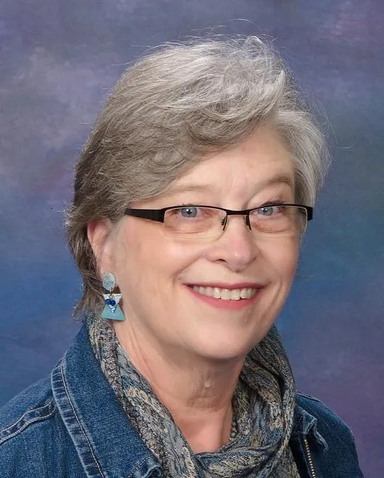 Headshot of author Carol Fitzpatrick smiling at the camera -- older woman with short, graying hair swept across the forehead, wire-rimmed glasses, a broad warm smile, and small turquoise earrings dangling from her lobes. She has a denim jacket and multi-hued scarf around her neck in hades of blue with a backdrop of soft blues and purples.