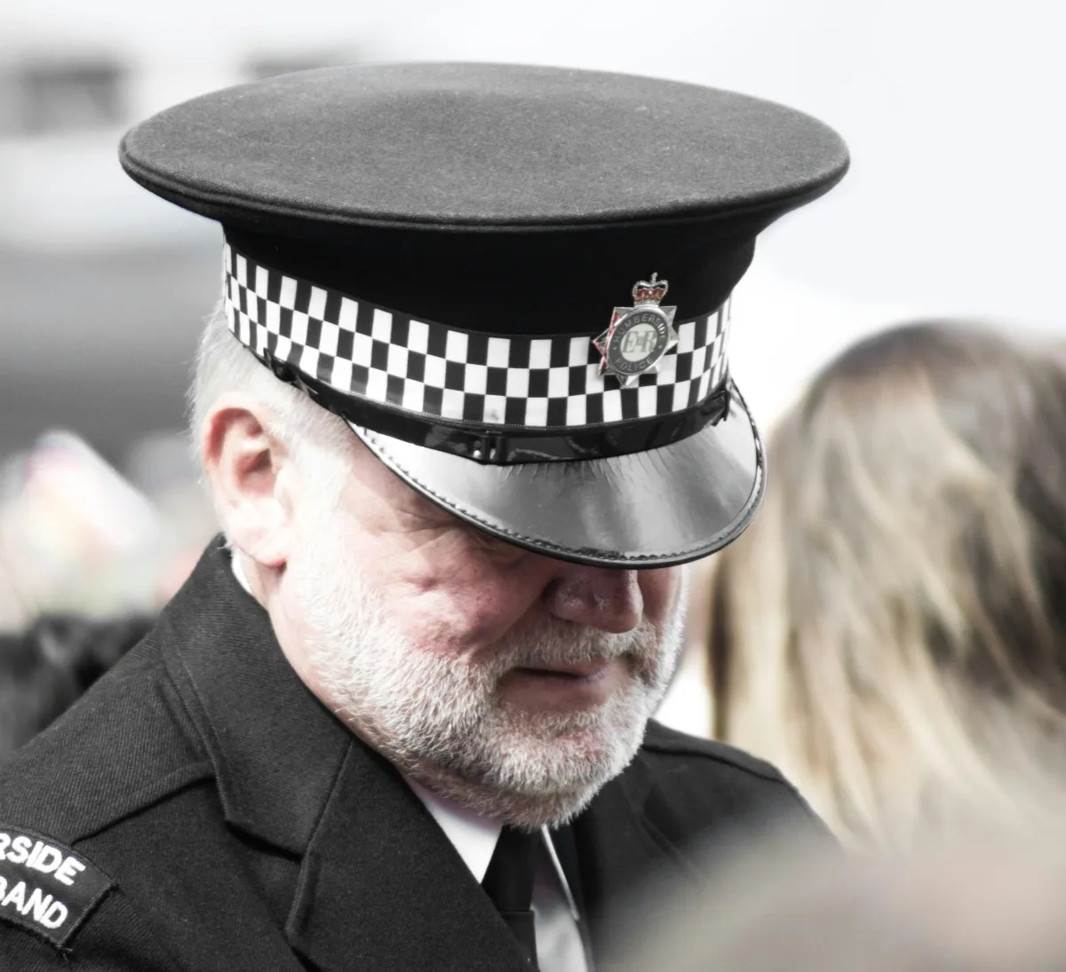 Officer Horace Nomos dressed in his formal officer garb cap pulled low over his face as he gazes downward, whitened beard shapely and trimmed about a solemn but kind mouth.