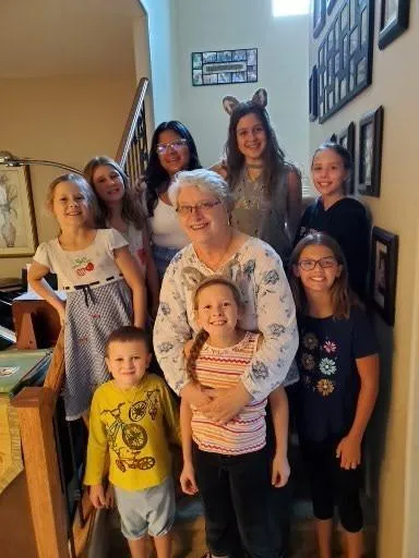 Photoraph of author Carol Fitzpatrick standing while embracing one of her beaming granddaughters while surrounded by seven other children (some are her grandchildren) all smiling at the camera in everyday outfits and looking delighted to be together for the photo.