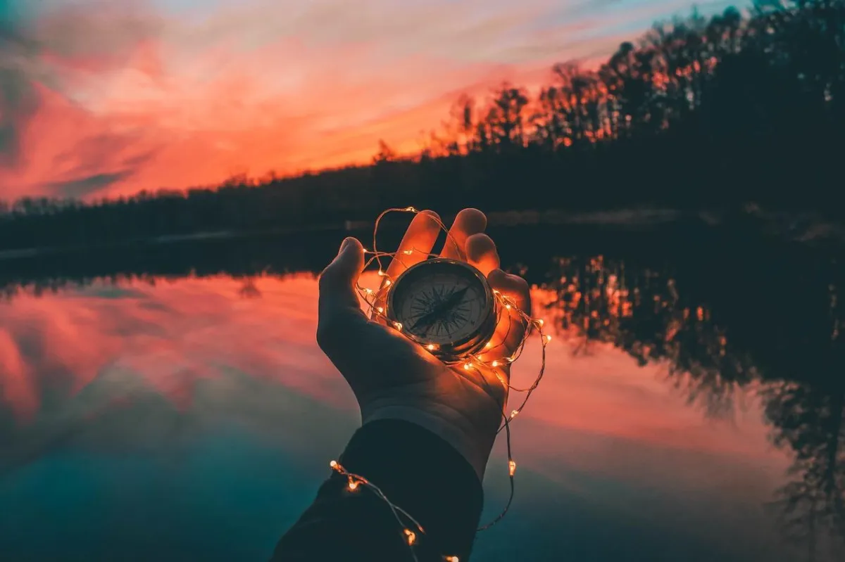 Compass held aloft in the palm of a hand at sunset over a lake and with the silhouette of woodlands on the farside