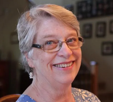 Headshot of author Carol Fitzpatrick smiling at the camera -- older woman with short, graying hair swept across the forehead, wire-rimmed glasses, a broad warm smile, and small coral earrings dangling from her lobes.