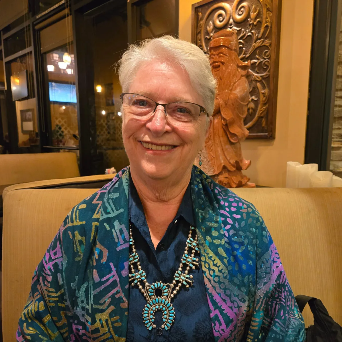Headshot of author Carol Fitzpatrick smiling at the camera -- older woman with short, graying hair swept across the forehead, wire-rimmed glasses, a broad warm smile, and wearing a colorful dark teal shirt and turquoise necklace
