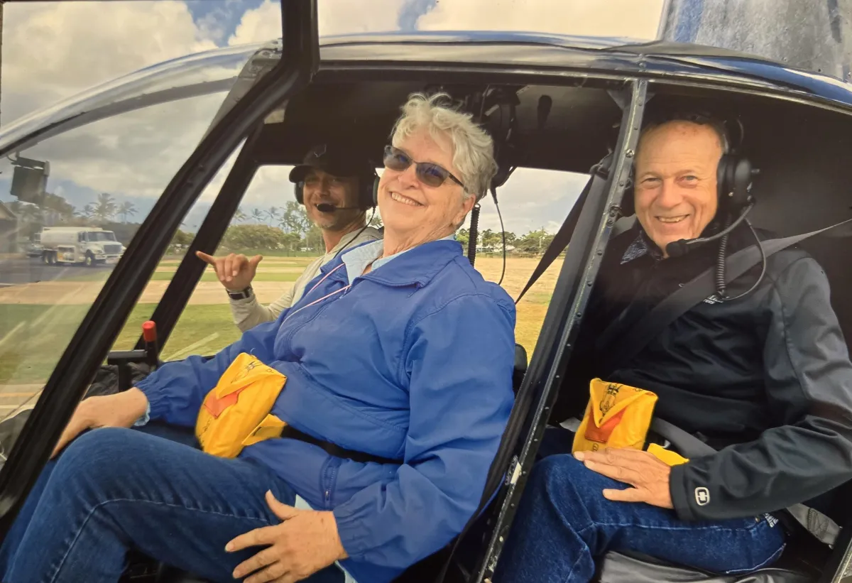 C. S. Fitzpatrick beaming from the front seat of an open helicopter cockpit with her husband donning headphones and grinning in the backseat, and the pilot throwing a shakaa as they prepare for a helicopter lesson in Kauai