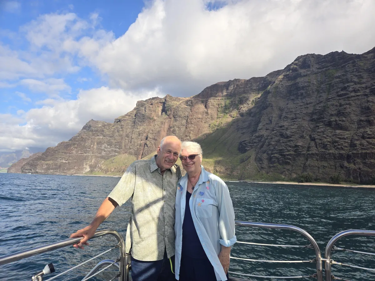 Leaning against a railing with her husband, C. S. Fitzpatrick stands smiling on the Na-Pali Coast, Kauai with large puffy clouds in a blue sky with rocky, reddish cliffs, and blue and teal waters behind