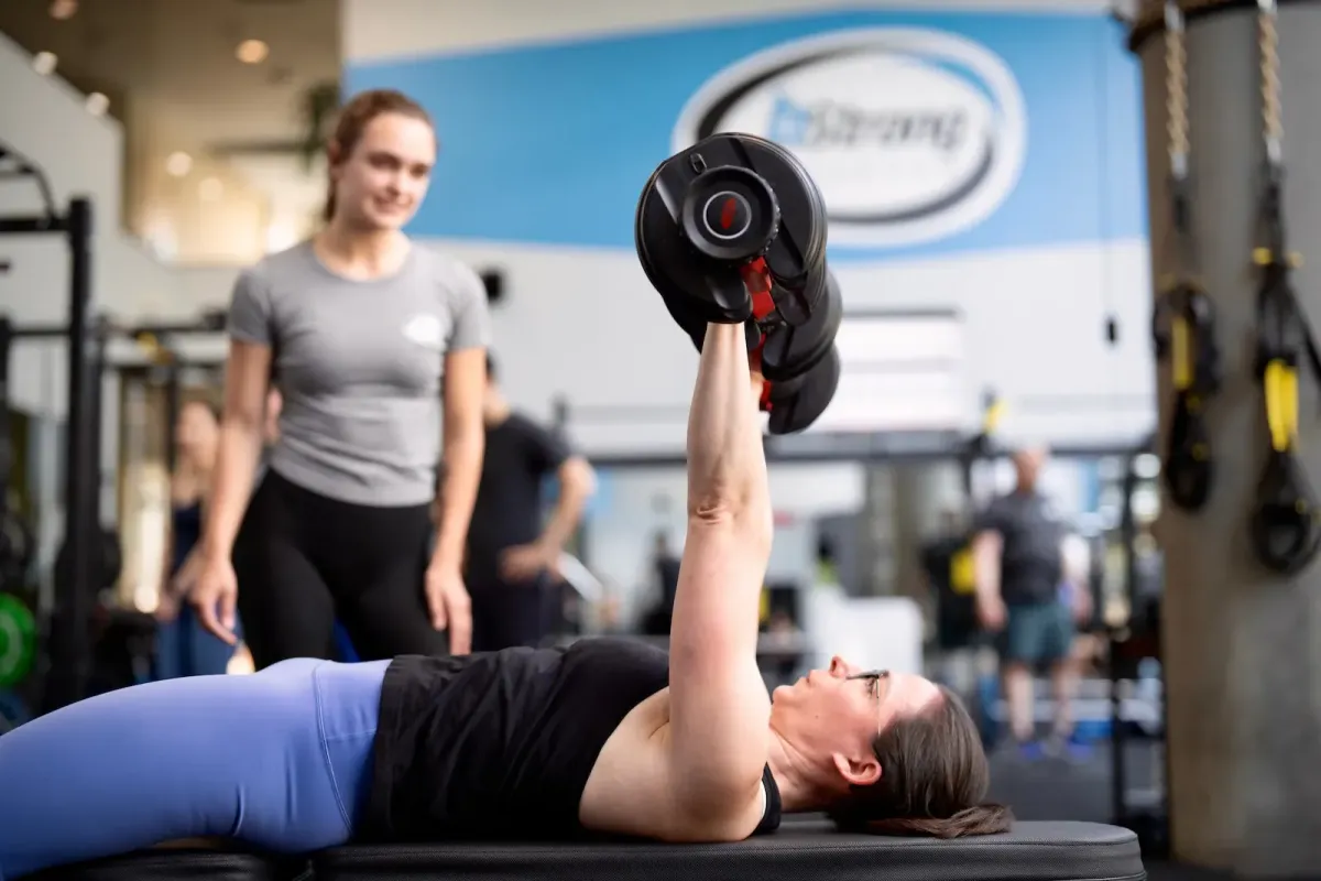 A member on a bench performing a chest press with a personal training looking on and guiding her. 