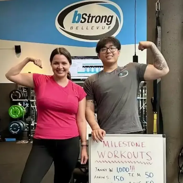 A member and a coach flexing in the gym with a sign celebrating a milestone workout for her. 