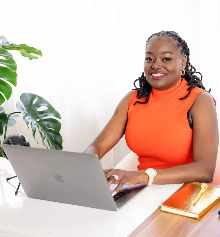 A woman wearing an orange top sits at her desk, focused on her work with a laptop in front of her.