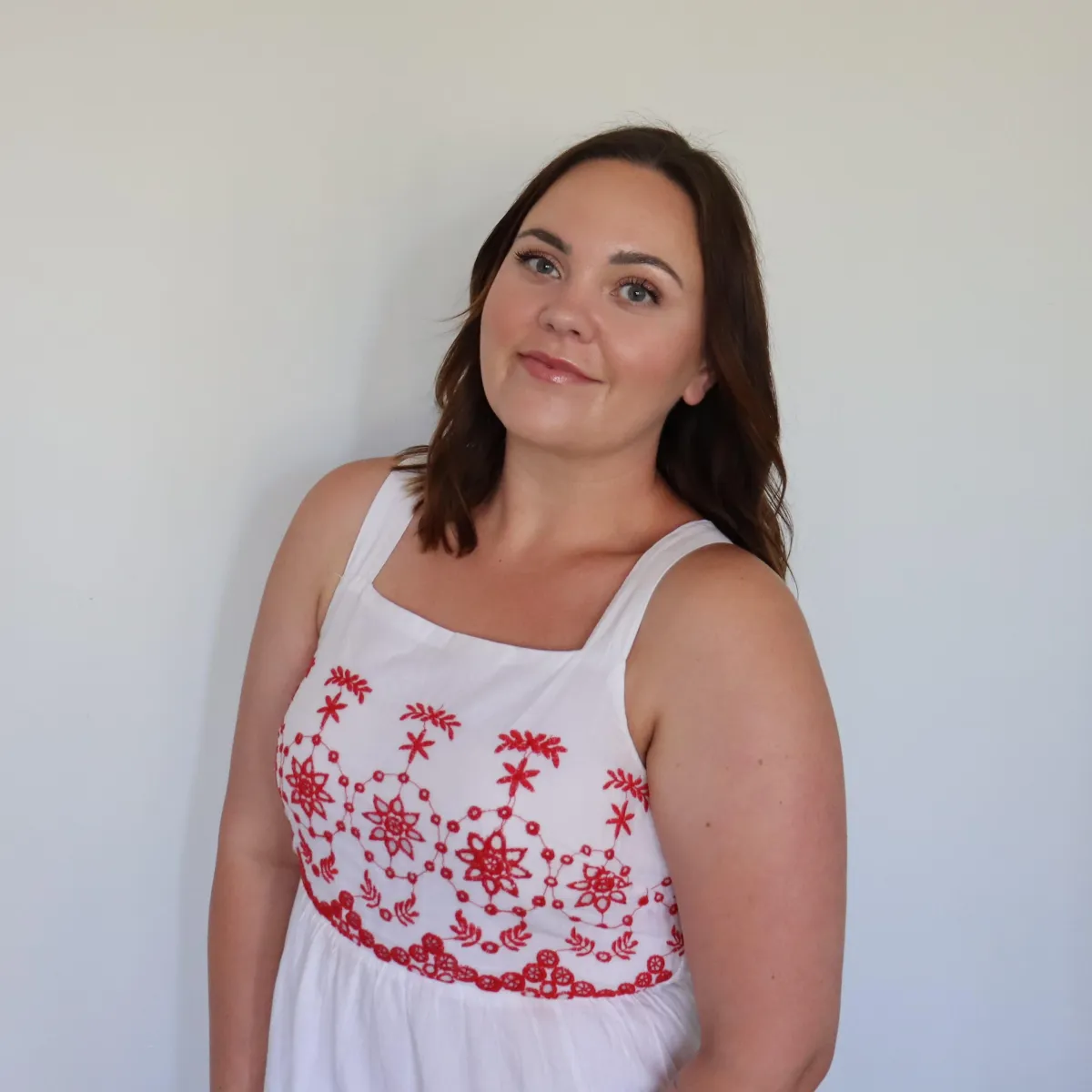 Woman in a white dress with red embroidery, smiling against a neutral background, representing approachable expertise in buyer advocacy at Arin Russell Buyers Agents.
