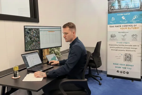 Person working at a desk with multiple monitors displaying property data and charts, featuring an Arin Russell Buyers Agents banner emphasizing property investment support and management.