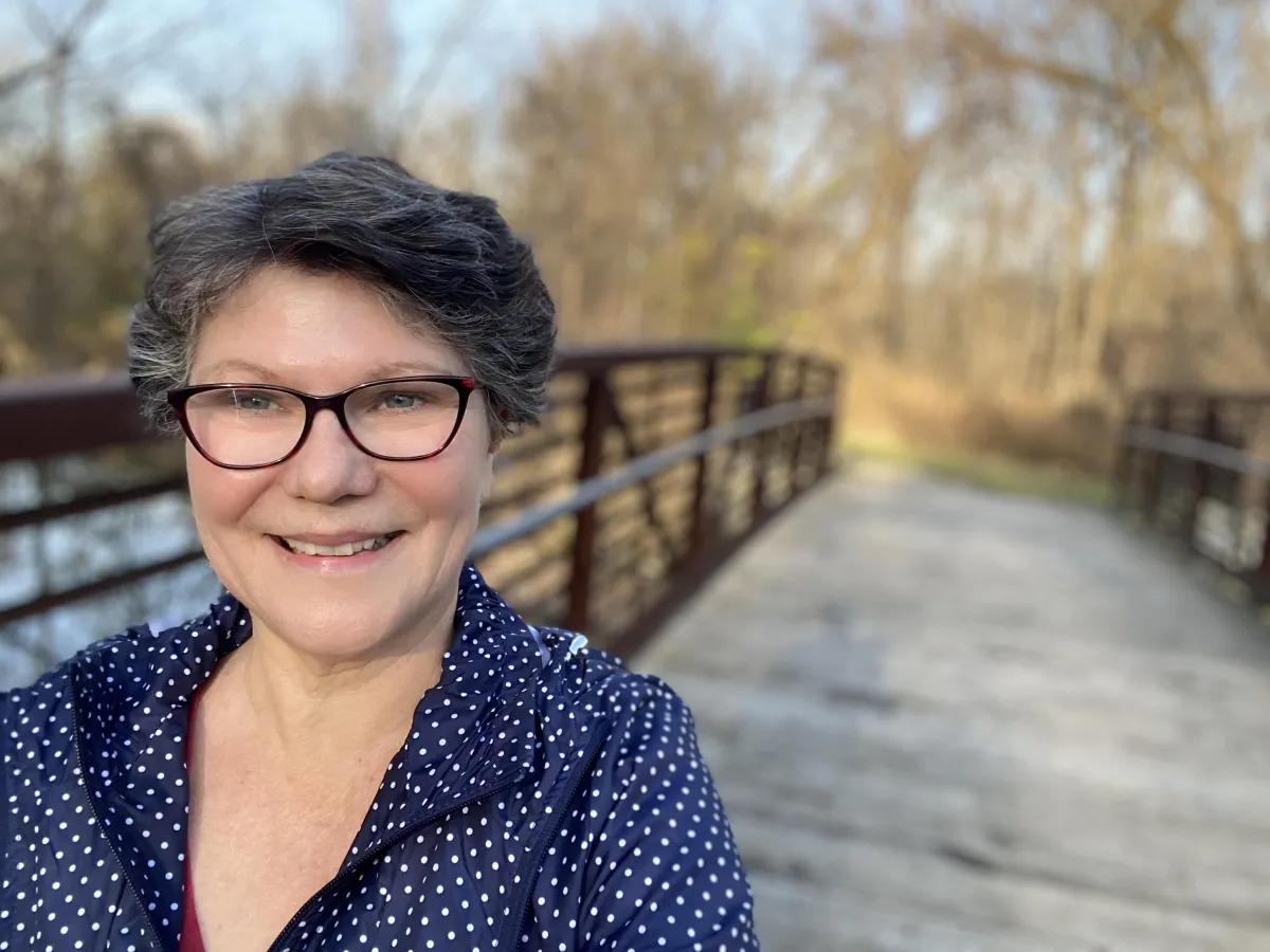 Janet standing outdoors on a wooden bridge, wearing a navy blue polka dot jacket. The background features trees and a serene natural setting with soft sunlight, creating a calm atmosphere. Perfect for done-for-you marketing automation services, highlighting approachability, trust, and personal connection.