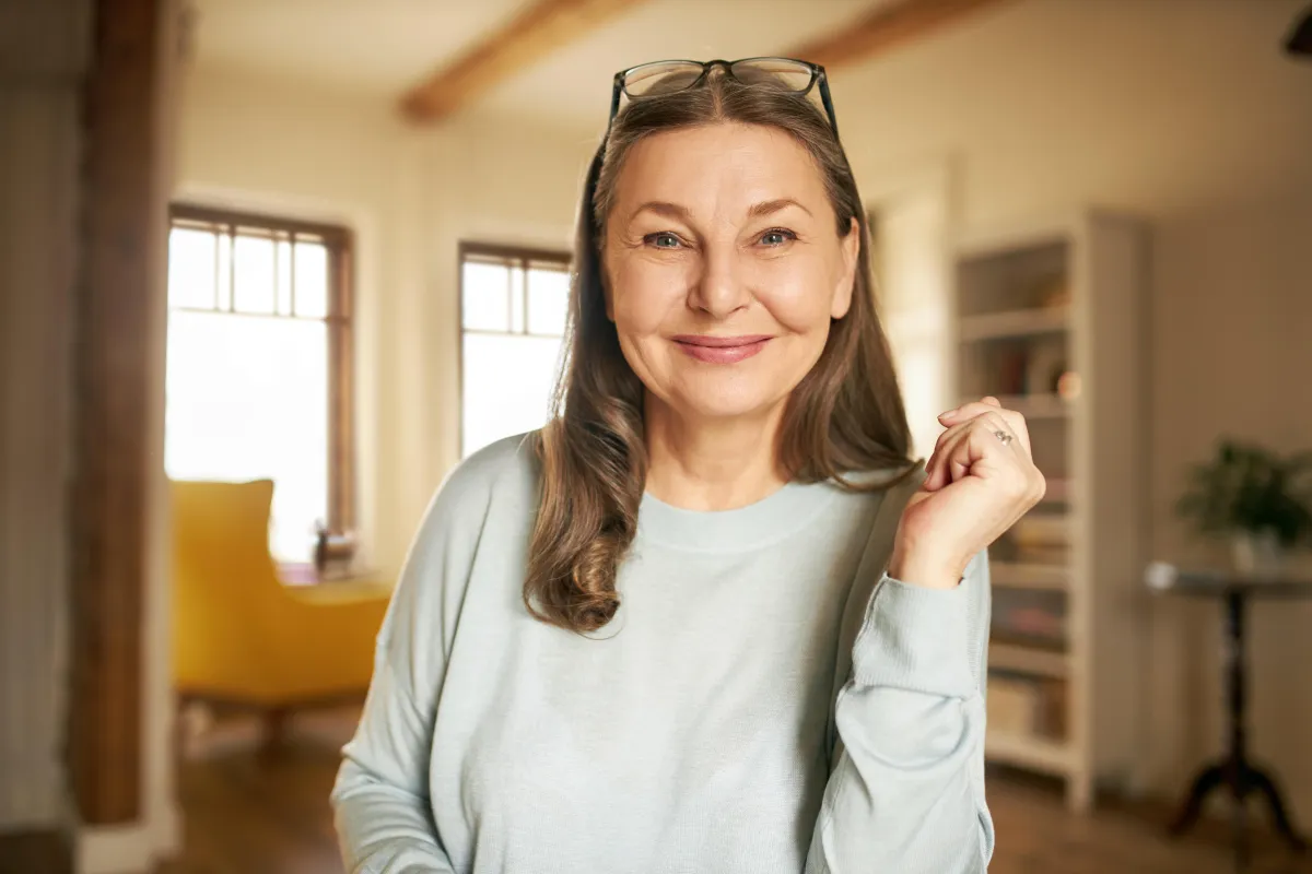  Professional woman with long brown hair, wearing glasses and a light grey sweater, sits in a cozy, sunlit home office with bookshelves and a yellow chair in the background. She smiles confidently, embodying ease and approachability. Ideal image for "60-Days to stress free, automated business. No tech skills required." — marketing automation services that require no technical expertise.