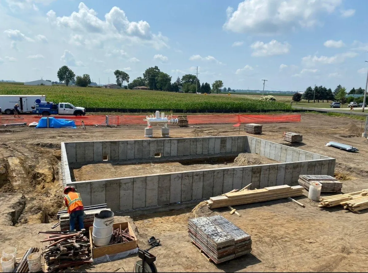 Construction site featuring a concrete foundation being prepared, with a worker in safety gear, construction materials, and machinery in a rural setting.