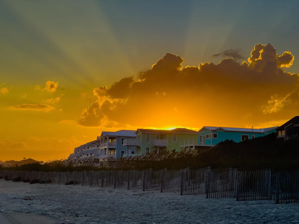 Vibrant sunset over beachfront houses, with rays of sunlight streaming through clouds, casting a golden glow across the sky and landscape. The image captures the serene beauty of coastal living, showcasing Drake Nix’s skill in sunset and landscape photography.