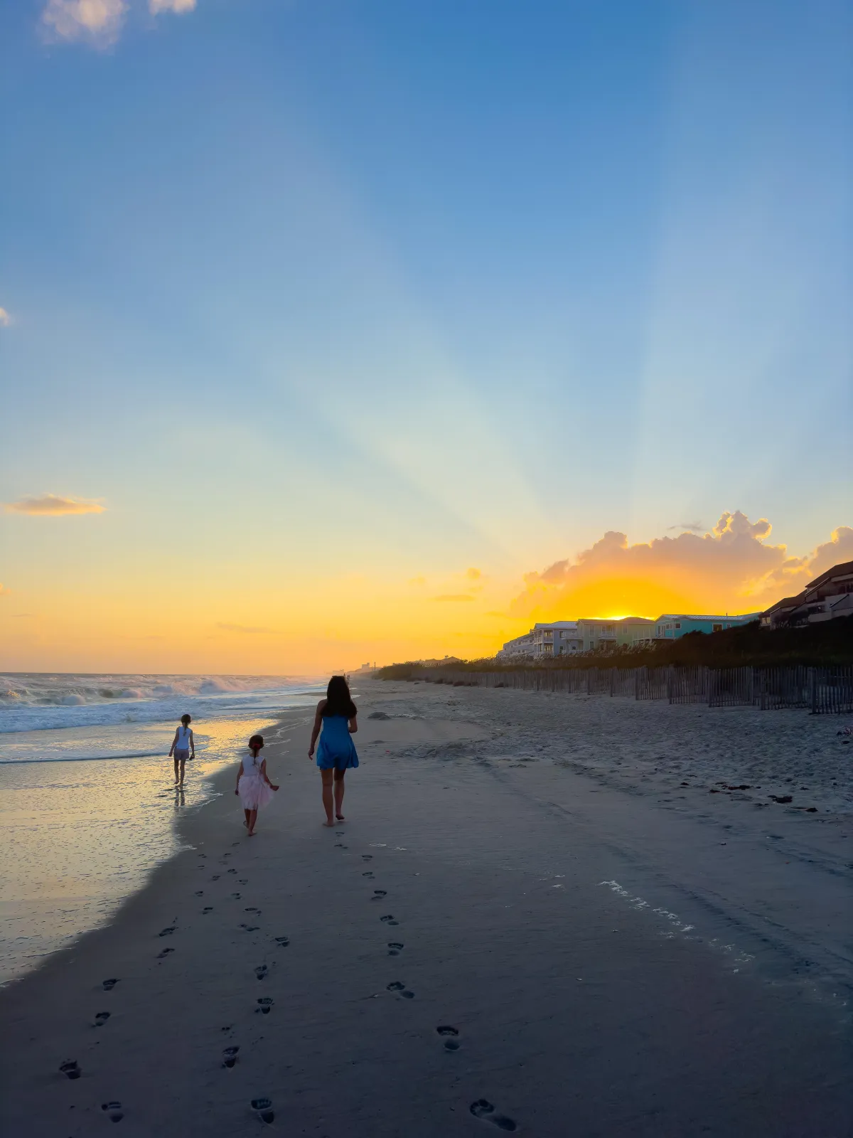 Sunset beach scene with a woman and two children walking along the shoreline, leaving footprints in the sand. Sun rays fan out from behind clouds, creating a serene and golden glow, capturing a peaceful family moment and highlighting Drake Nix’s talent in lifestyle and sunset photography.