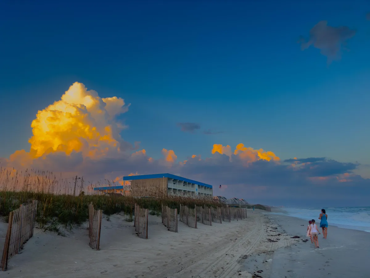 Serene beach scene at sunset with golden clouds towering over the horizon, a building nestled by dune fences, and a family walking along the shore. The image captures a peaceful coastal atmosphere, highlighting Drake Nix’s talent in landscape and lifestyle photography.