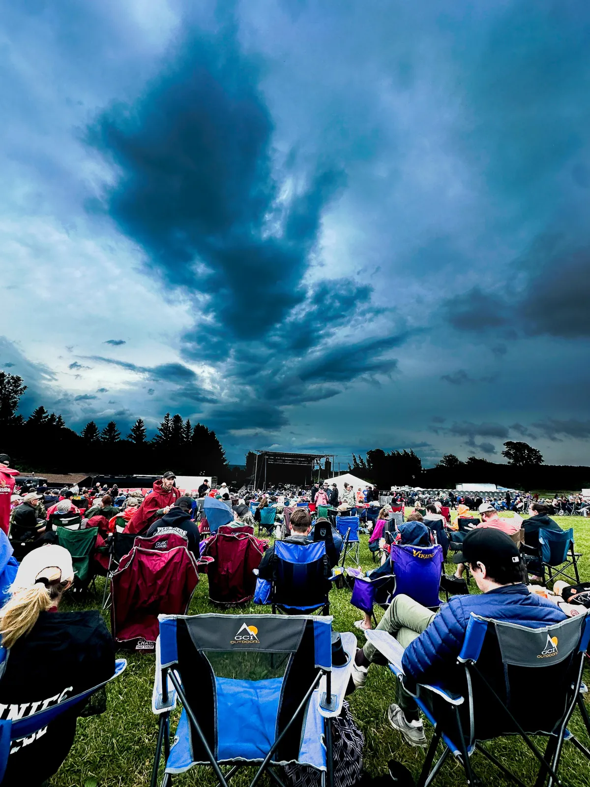 Outdoor concert scene with an expansive, dramatic sky and an audience seated in colorful camping chairs facing a distant stage. The image captures the atmosphere of a live event under dynamic cloud formations, showcasing Drake Nix’s skill in event photography and capturing mood.