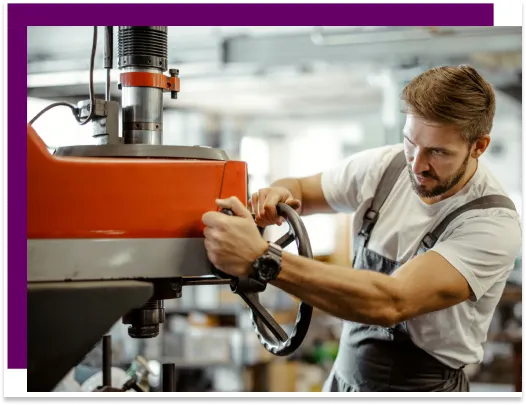 Konzentrierter Industriearbeiter mit Bart und Arbeitskleidung bedient eine große rote Werkzeugmaschine in einer gut ausgestatteten Werkstatt, was handwerkliches Können und Fachwissen im Maschinenbau illustriert.