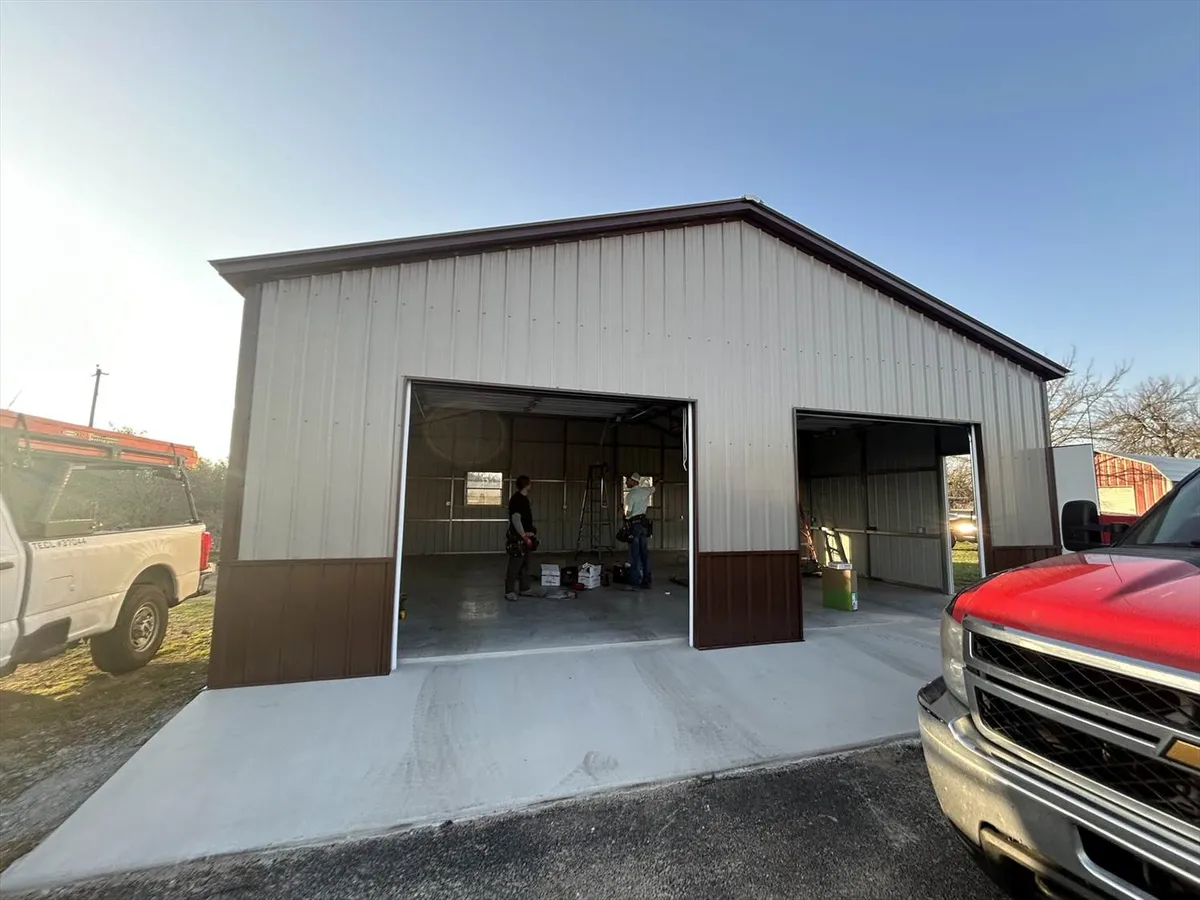 A white and brown metal barn or workshop building with open bay doors revealing the interior, a red truck parked outside.