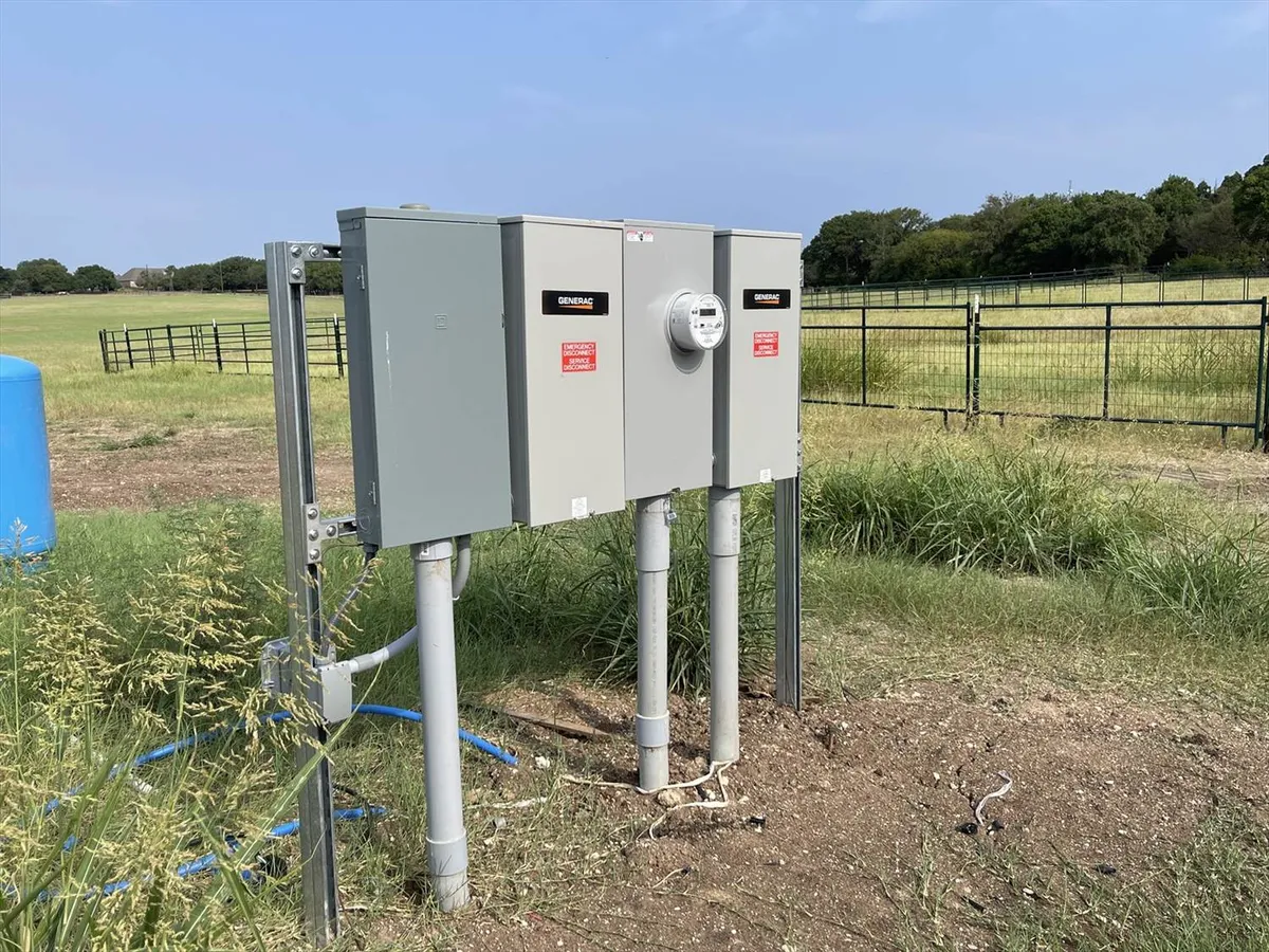 Outdoor electrical utility meters and breaker boxes mounted on metal stands in a grassy field with a fence in the background.