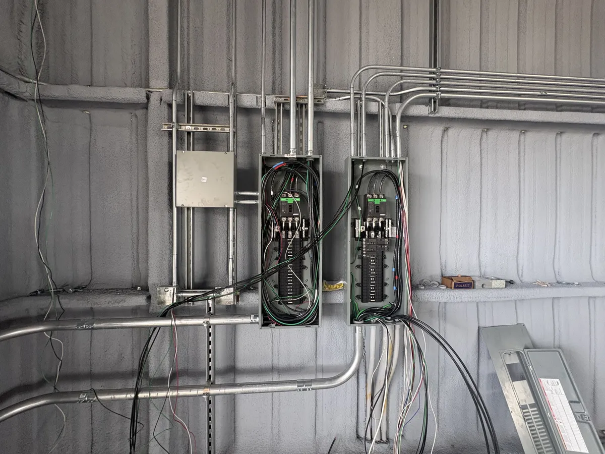 Multiple gray electrical breaker panels and metallic conduit pipes installed on a silver corrugated metal interior wall, typical of a warehouse or commercial building