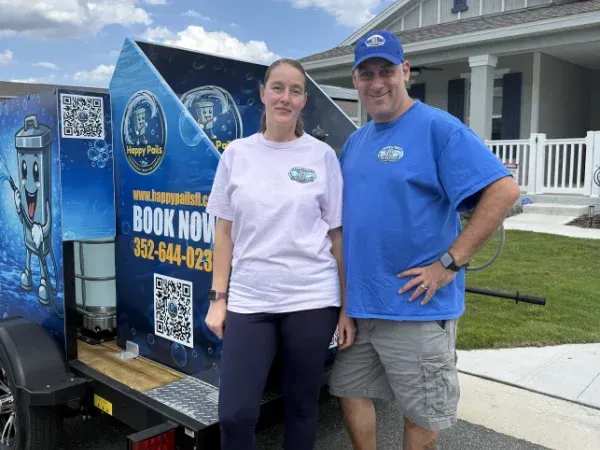 A man and woman smiling in front of a Happy Pails pressure washing trailer with branding, during daytime.
