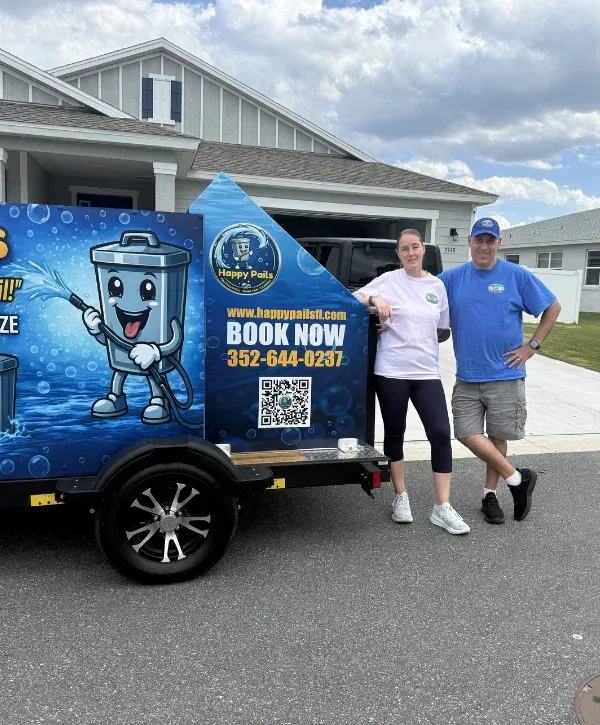 A man and woman standing next to a black utility trailer with a blue Happy Pails trash can cleaning advertisement.