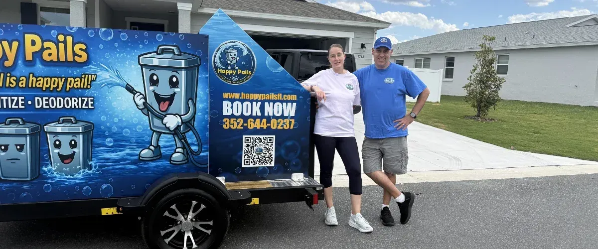 A man and woman standing next to a black utility trailer with a blue Happy Pails trash can cleaning advertisement.