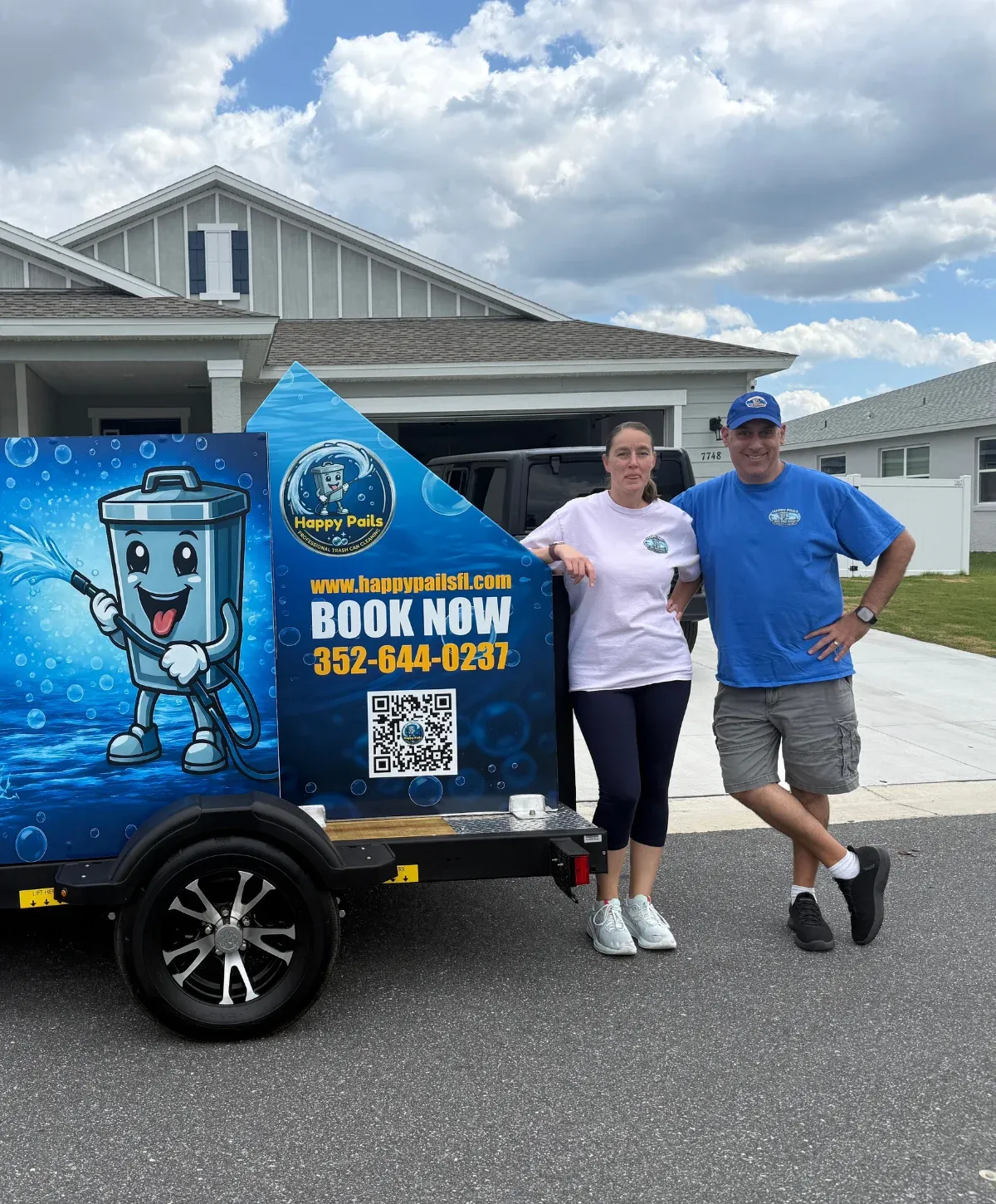 Owners of Happy Pails Professional Trash Can Cleaning stand proudly beside their branded cleaning trailer with a cartoon trash can logo.
